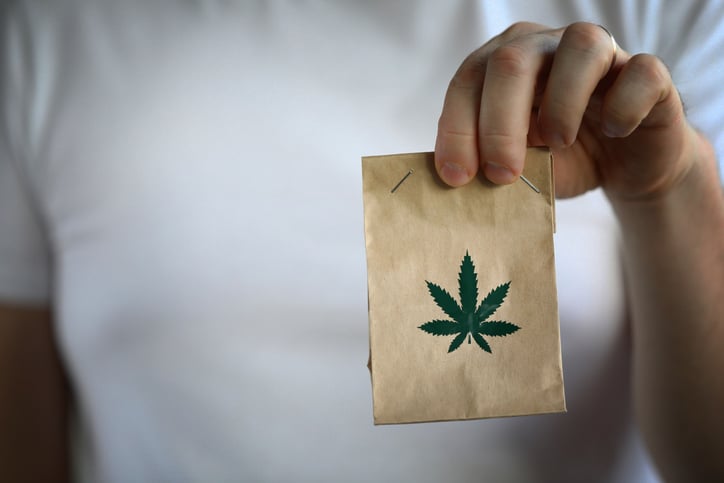 A person holding a small brown paper bag with a marijuana leaf graphic on it.